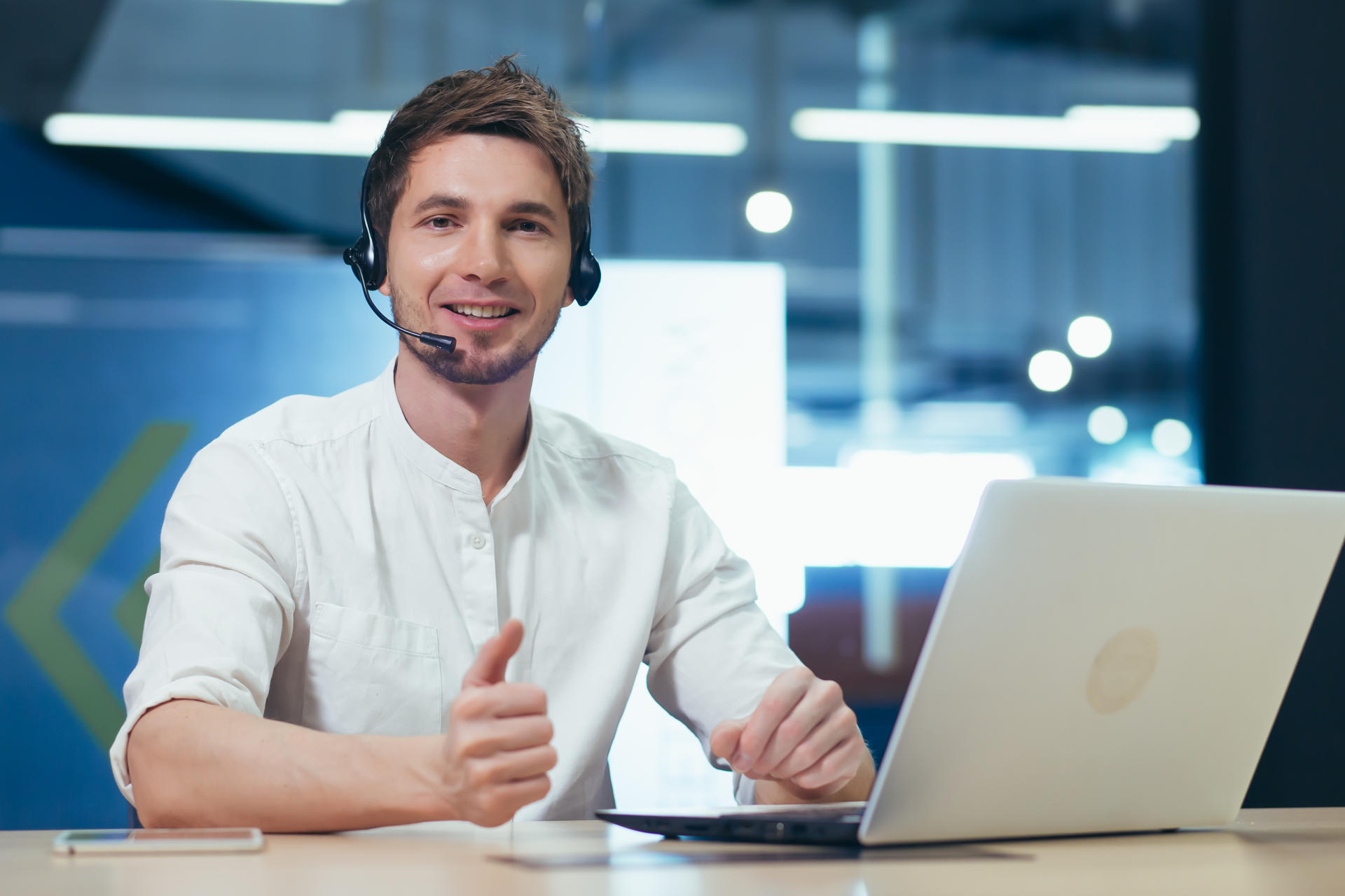 Portrait of a young call center operator, a man smiling and looking at the camera, working with a laptop using a headset for a video call